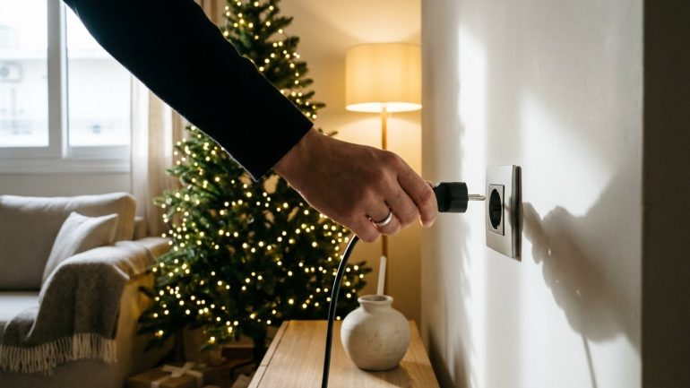 Χέρι που βγάζει από την πρίζα τα λαμπάκια του χριστουγεννιάτικου δέντρου σε φωτεινό σαλόνι.