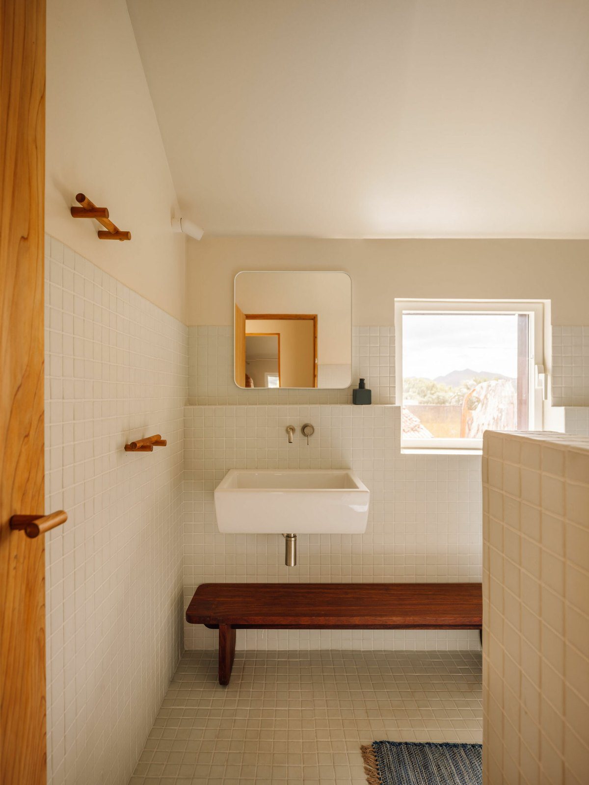 Μπάνιο με λευκό νιπτήρα τοίχου, στρογγυλεμένο καθρέφτη, ξύλινο πάγκο και ξύλινες κρεμάστρες στον τοίχο