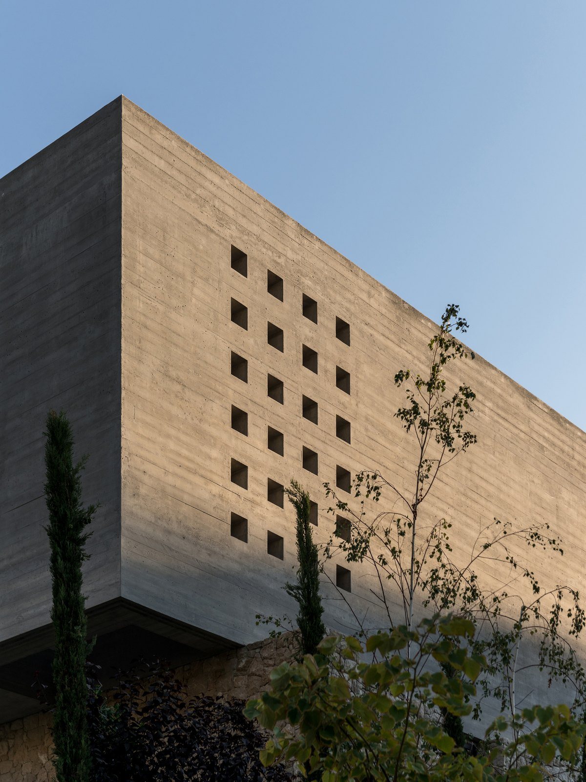 Τοίχος από εμφανές μπετόν με κάνναβο από μικρά τετράγωνα ανοίγματα και κυπαρίσσια μπροστά του