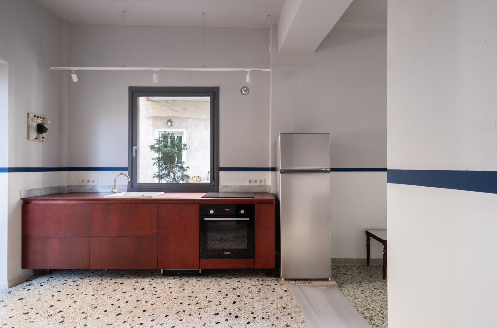 Κουζίνα σε ευθεία με κόκκινα ξύλινα ντουλάπια, παράθυρο πάνω από τον νεροχύτη, ψυγείο και μωσαϊκό δάπεδο, με μπλε λωρίδα στους λευκούς τοίχους