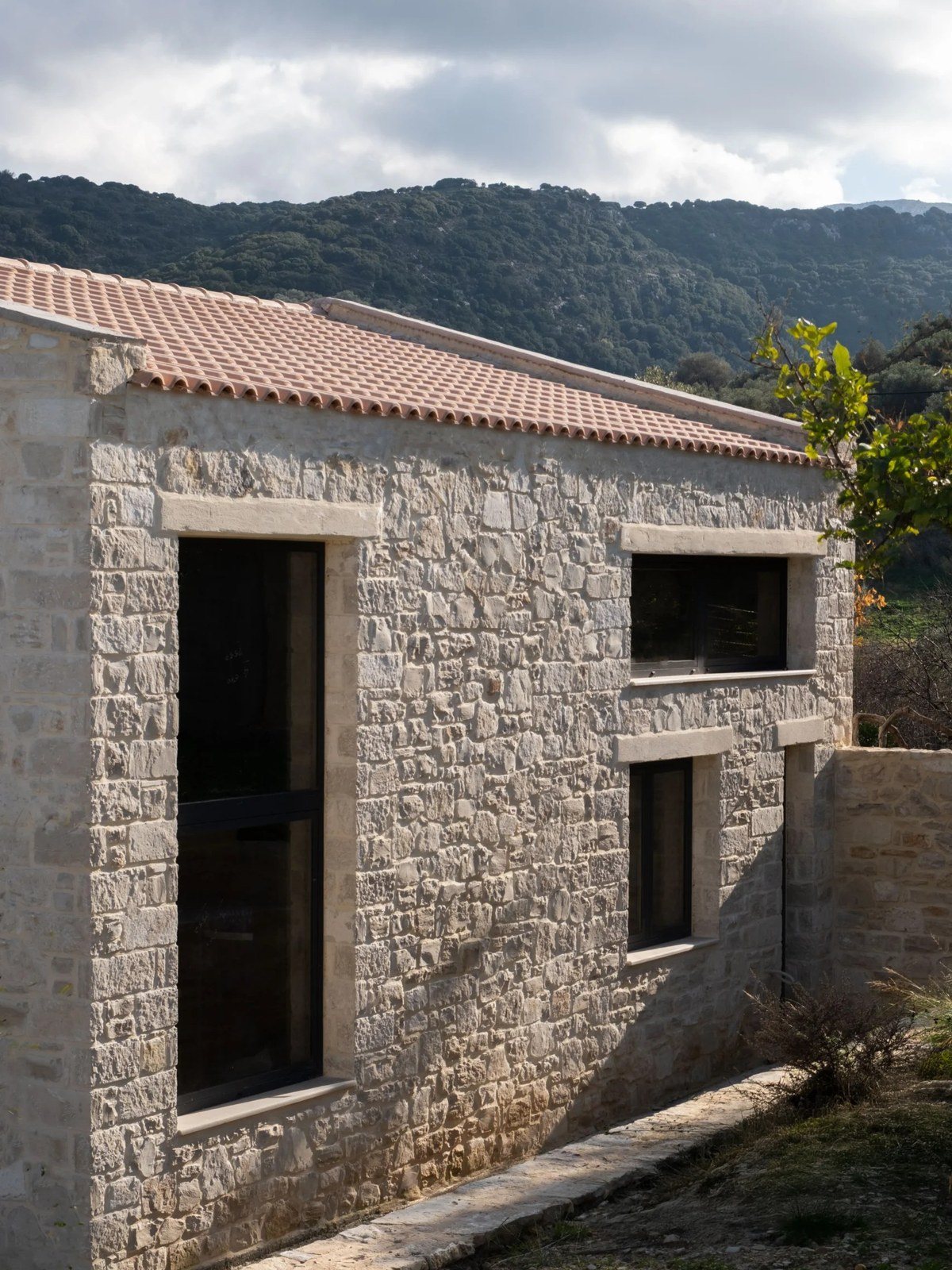 Πλάγια όψη πέτρινου σπιτιού με κεραμοσκεπή, ψηλό στενό παράθυρο και δύο μικρότερα, με πράσινο βουνό στο βάθος