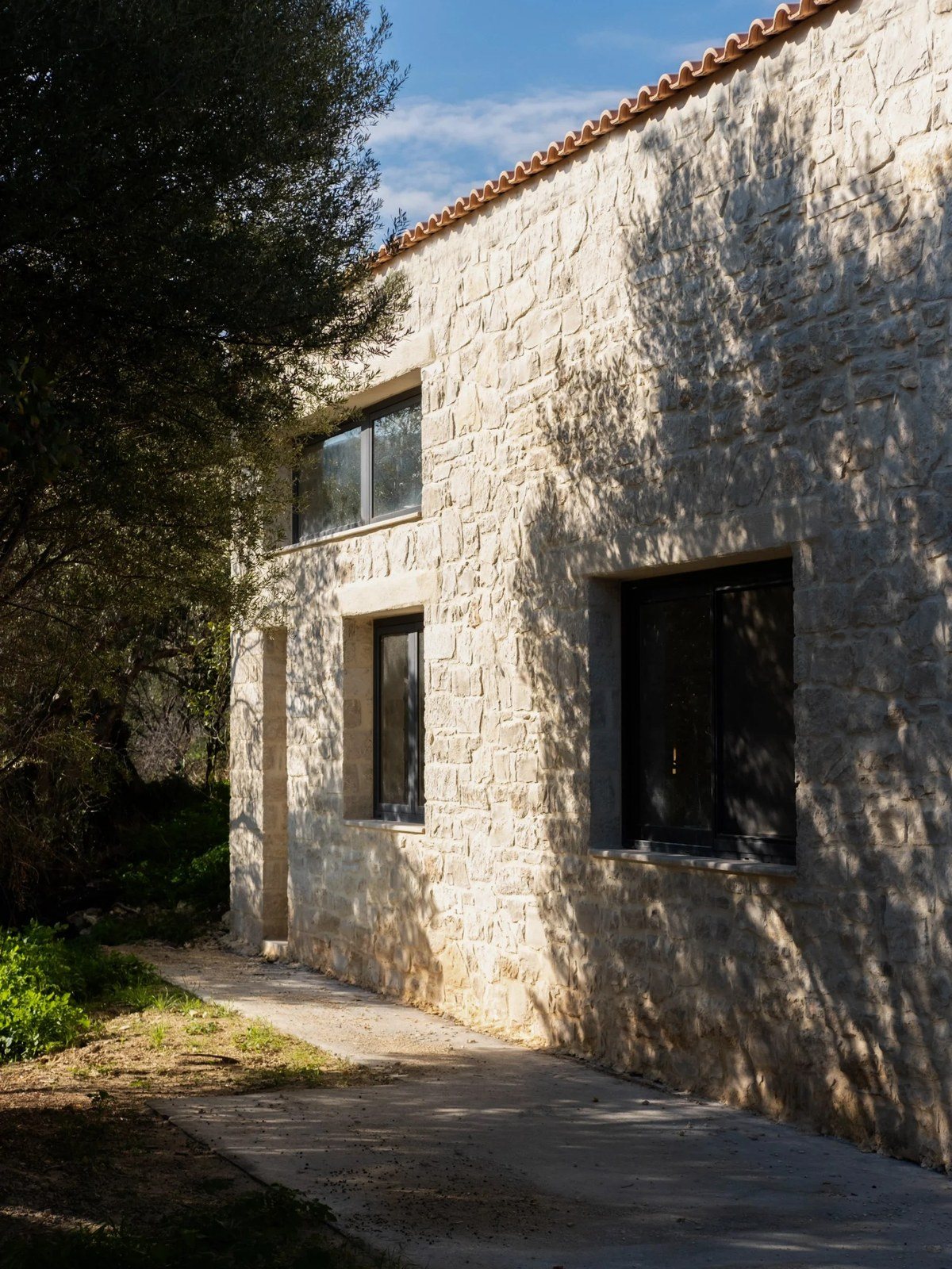 Πέτρινη όψη με κεραμοσκεπή και σκούρα παράθυρα, με σκιές από κλαδιά ελιάς και μονοπάτι δίπλα στον τοίχο