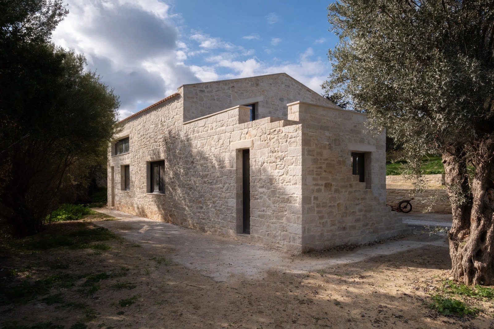 Πέτρινο σπίτι με μικρά σκούρα παράθυρα και κλιμακωτό στηθαίο ανάμεσα σε δύο μεγάλες ελιές, πάνω σε χωμάτινο έδαφος