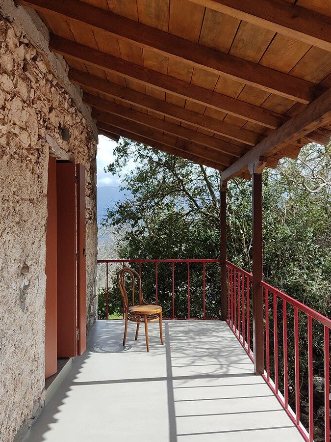 Στενή βεράντα με κόκκινα κάγκελα, ξύλινη στέγη, πέτρινο τοίχο και μια ξύλινη καρέκλα με θέα σε δέντρα