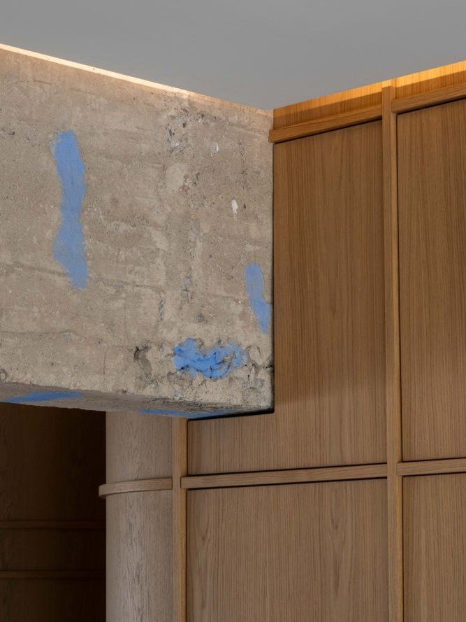 Γυμνό δοκάρι από μπετόν με μπλε σημάδια που συναντά ξύλινα πάνελ δρυός με κρυφό φωτισμό στο ταβάνι