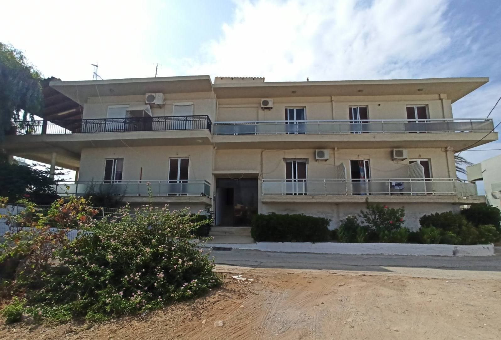 Τριώροφο ξενοδοχείο με μπεζ όψη, μακριά μπαλκόνια με κάγκελα και κλιματιστικά, πίσω από χωματόδρομο και θάμνους