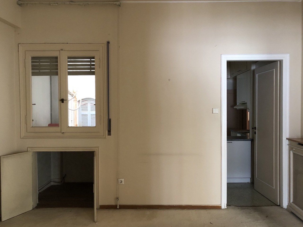 Παλιός τοίχος με παράθυρο, χαμηλό άνοιγμα και πόρτα προς μια μικρή κλειστή κουζίνα, πριν από την ανακαίνιση