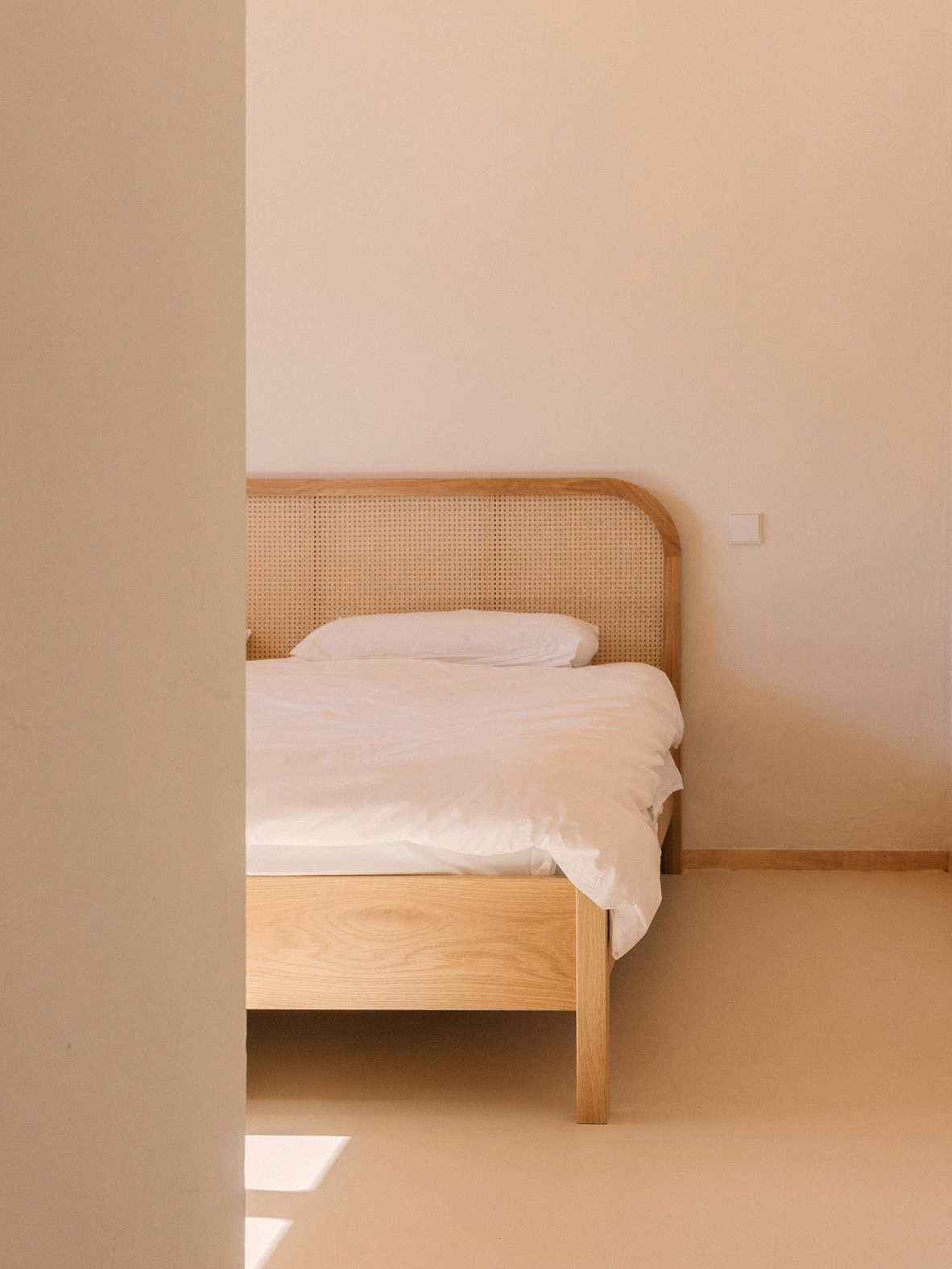 Λιτό υπνοδωμάτιο με ξύλινο κρεβάτι με ψάθινο κεφαλάρι, λευκά σεντόνια και ανοιχτόχρωμους τοίχους