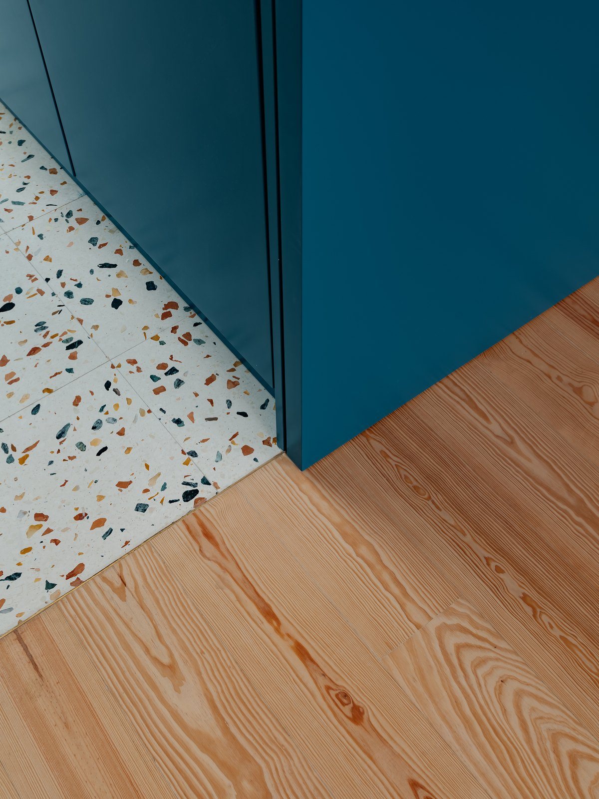 Δάπεδο terrazzo με χρωματιστά βότσαλα που συναντά διαγώνια ξύλινο πάτωμα από πεύκο στη γωνία του μπλε όγκου
