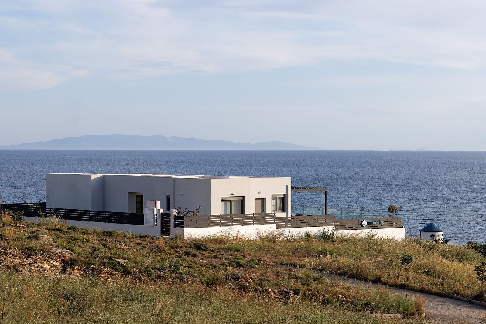 Λευκό λιτό κτίριο πάνω σε πλαγιά με ξερά χόρτα, με το Αιγαίο και ένα νησί στον ορίζοντα