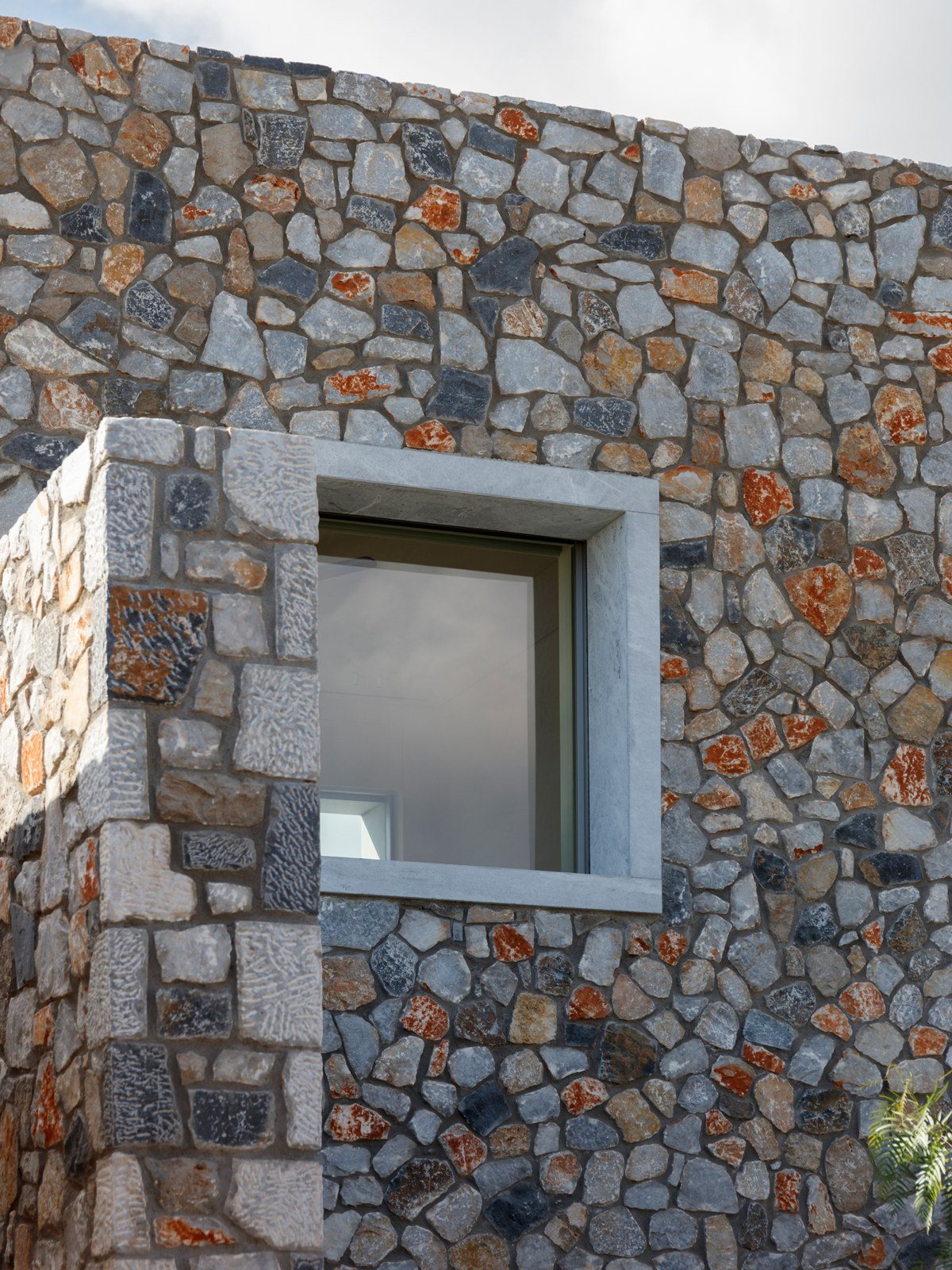 Λιθοδομή από ακανόνιστη πέτρα σε γκρι και σκουριά με τετράγωνο παράθυρο σε πλαίσιο από ανοιχτόχρωμο μάρμαρο