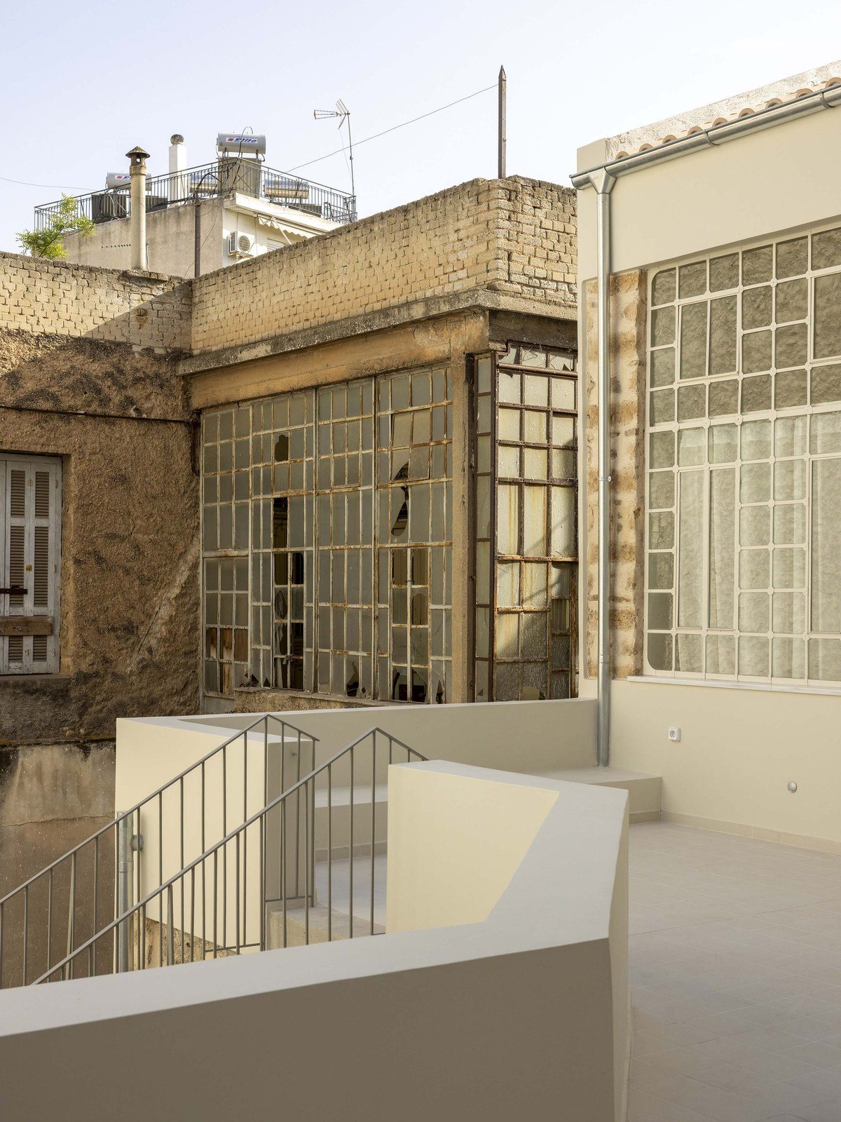 Βεράντα με λευκό στηθαίο και μεταλλικό κάγκελο σκάλας, ανάμεσα σε πέτρινους τοίχους και μεγάλα παράθυρα με μεταλλικά πλαίσια