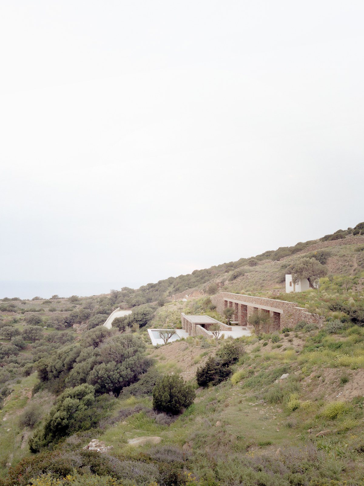 Πλαγιά με θάμνους και πεζούλες στη Σίφνο, όπου ένα σπίτι από πέτρα χάνεται μέσα στο έδαφος και ξεχωρίζει μόνο ένα μικρό λευκό κτίσμα